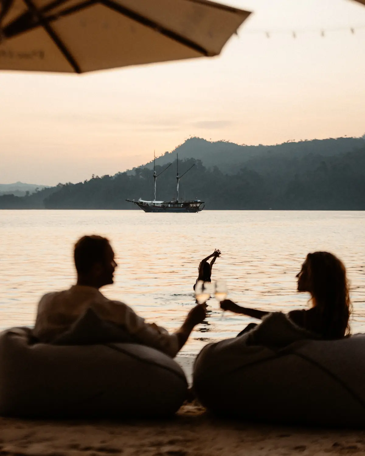 A couple on the beach at dusk with a phinisi anchored on the horizon