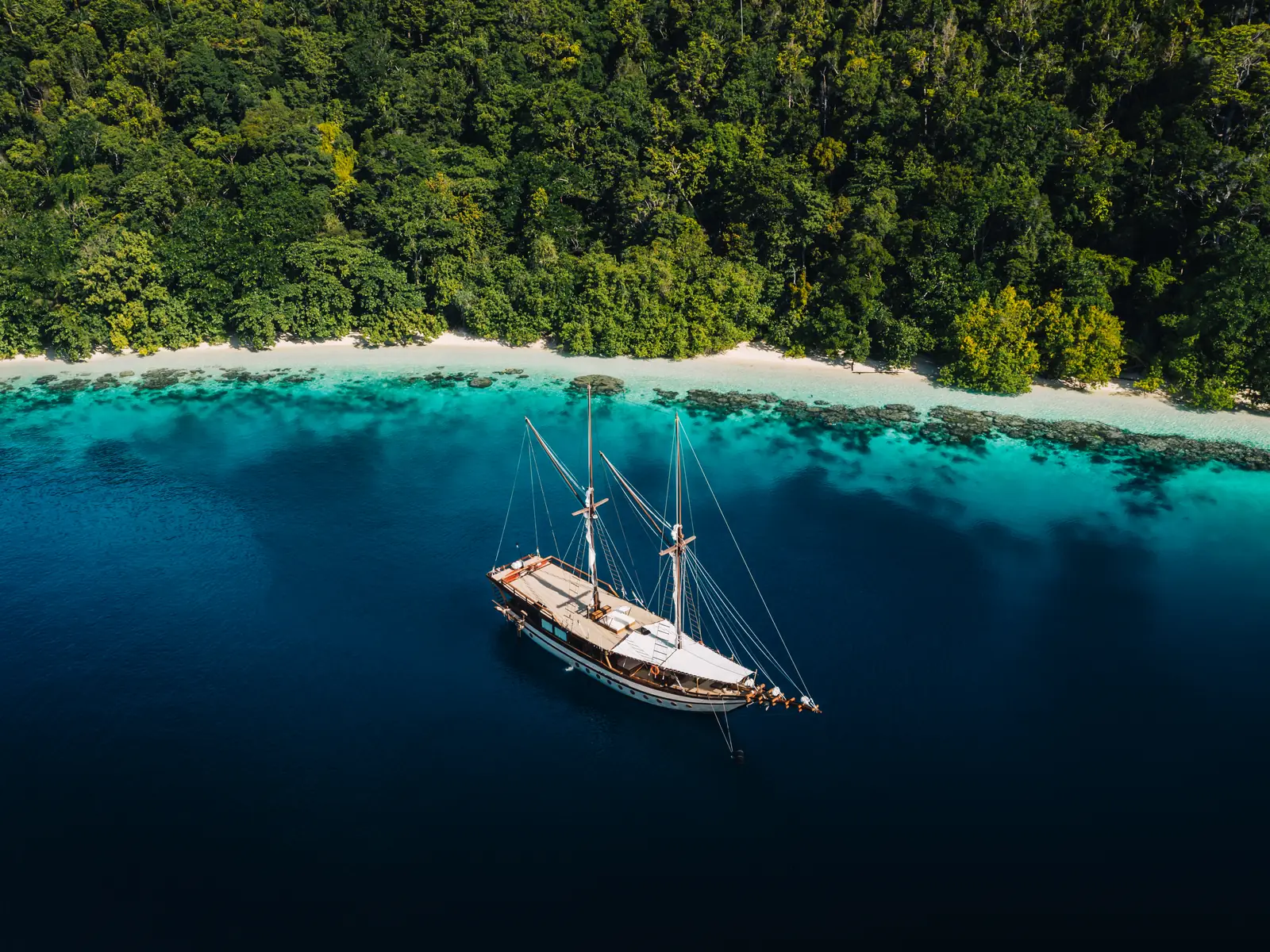 A phinisi anchored on the edge of a turquoise reef beside a white sand beach
